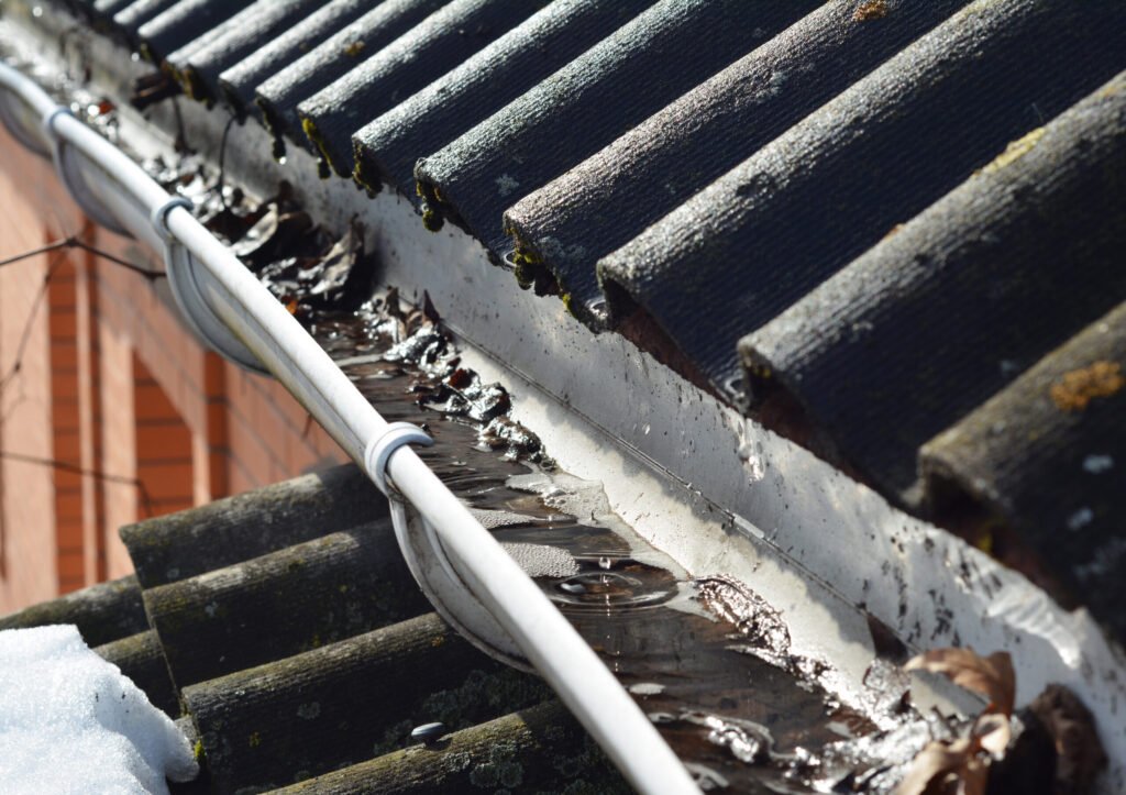 Roof gutter cleaning. A clogged rain gutter of an asbestos roof with non flowing water from melted snow because of unclean gutters with dirt and fallen leaves.