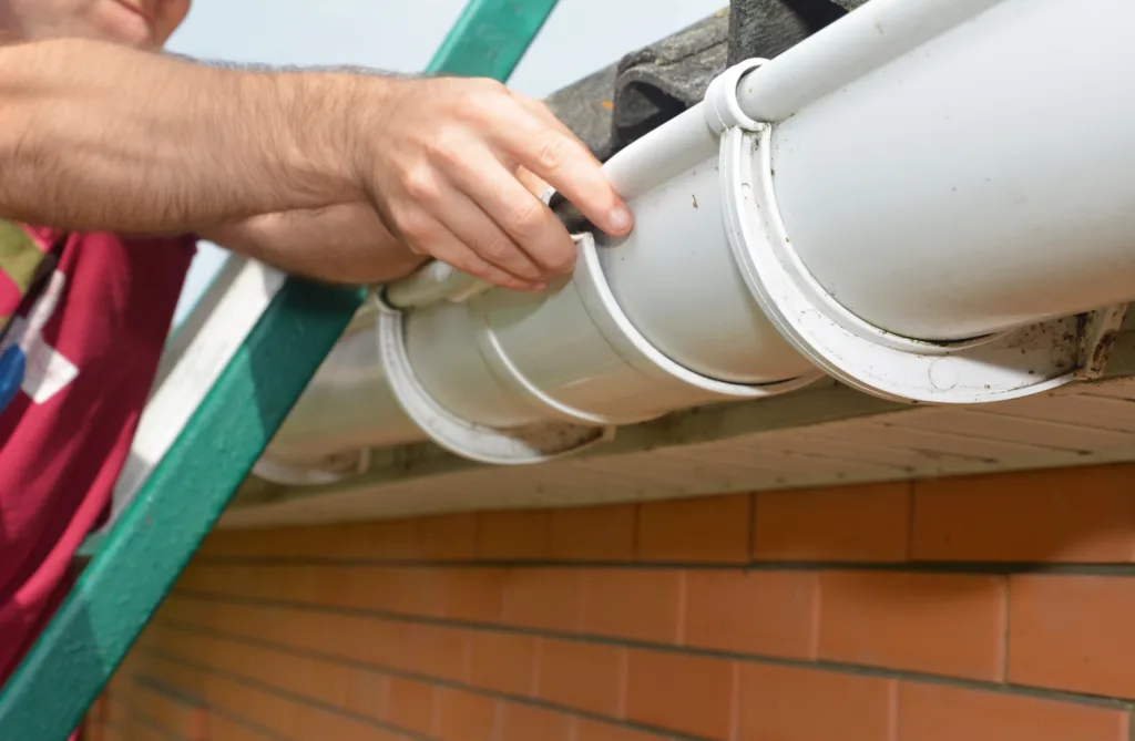 Roof gutter installation. A man on a ladder is installing a roof gutter’s slip joint connector to connect the gutters and avoid water leaks.